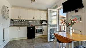 Eine kompakte Küche mit weißen Schränken, einem Geschirrspüler, einem Backofen und einer Mikrowelle. Ein Esstisch mit Stuhl, ein an der Wand befestigter Fernseher und ein Fenster mit Milchglas.