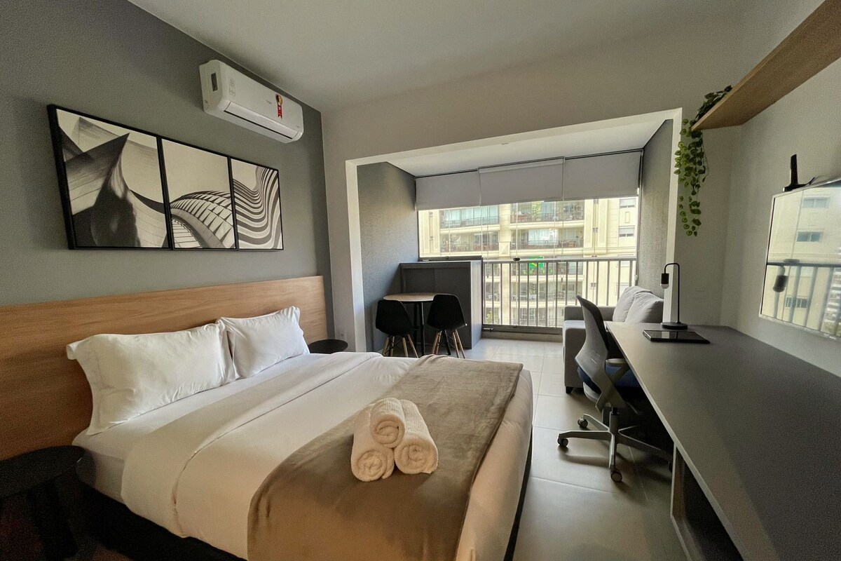 A hotel room with a bed, a desk, a chair, a balcony, and a view of buildings.