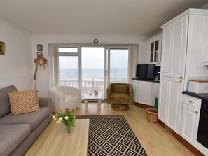 A living room with a sofa, a coffee table with a vase of flowers, a patterned rug, a television, and a kitchen area with cabinets and a countertop. - 1 Bed in Saundersfoot (oc-fb246) (Saundersfoot)