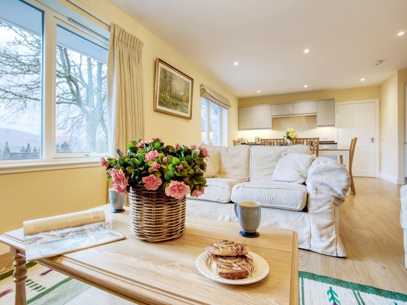 A cozy living room with a sofa, a dining table with a basket of flowers, and a view of the kitchen.