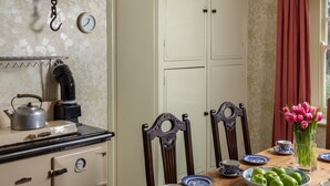 Eine Küche mit Herd, Wasserkocher und einer Uhr an der Wand. Ein Esstisch mit Stühlen, Tellern und einer Blumenvase steht bereit.