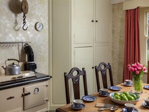 Cocina con estufa, hervidor y un reloj en la pared. Hay una mesa de comedor con sillas, platos y un florero con flores.