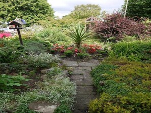 Un jardín con un sendero de piedra, un comedero para pájaros y diversas plantas.