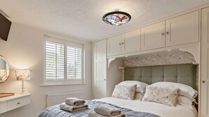 Ein Schlafzimmer mit Bett, Fenster mit Jalousien, Heizkörper, Nachttisch mit Lampe und Deckenleuchte.