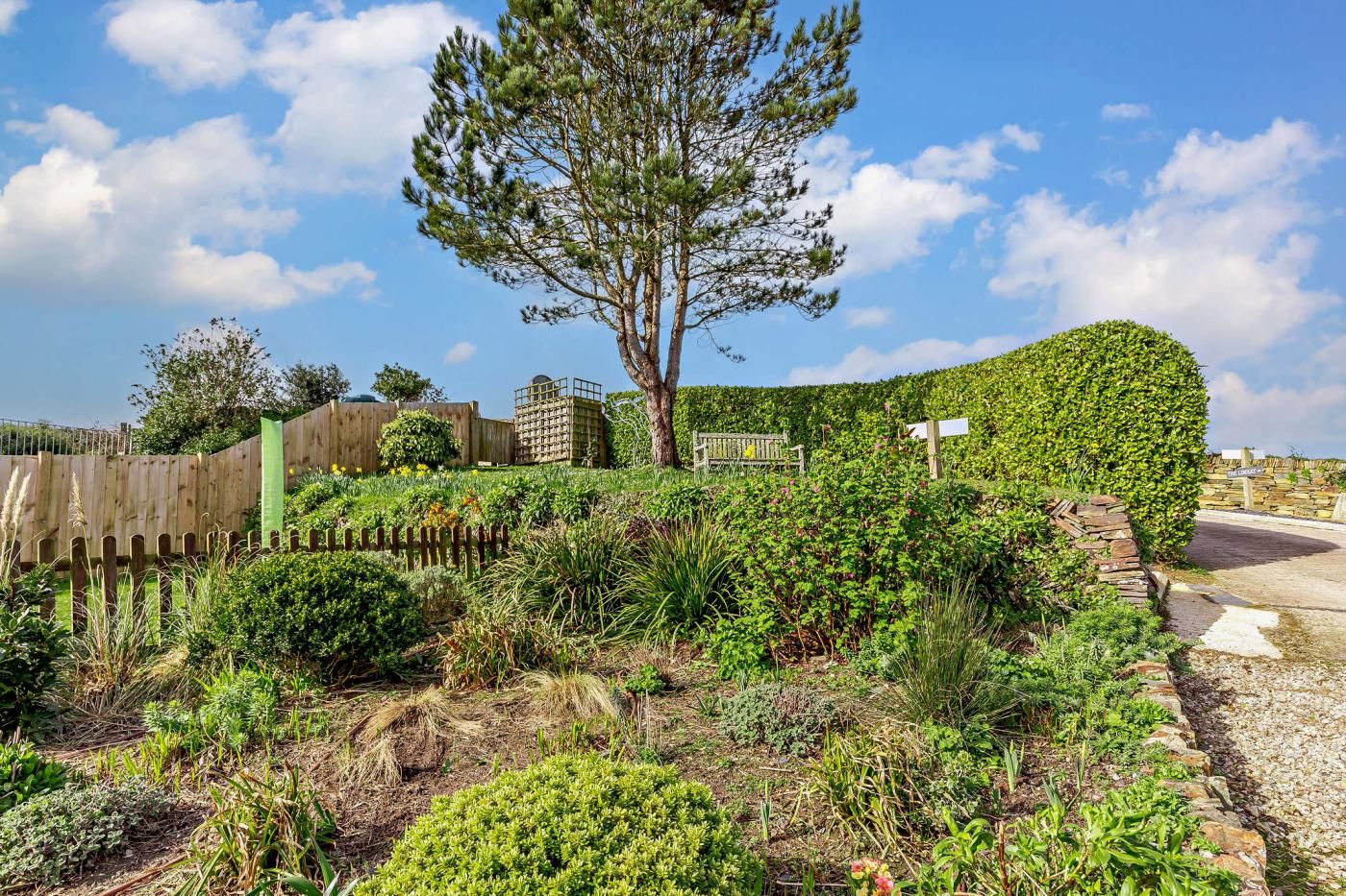 Un jardín con diversas plantas, una valla de madera, un banco y un cartel.