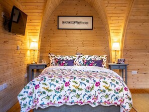 A bedroom with a sloped ceiling, wooden walls, a bed with floral bedding, two bedside tables with lamps, and a framed picture on the wall.