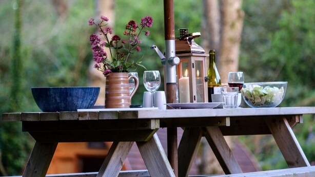 Een houten tafel buiten gedekt, met een kom, een gestreepte mok, een kaars, een fles en glazen.