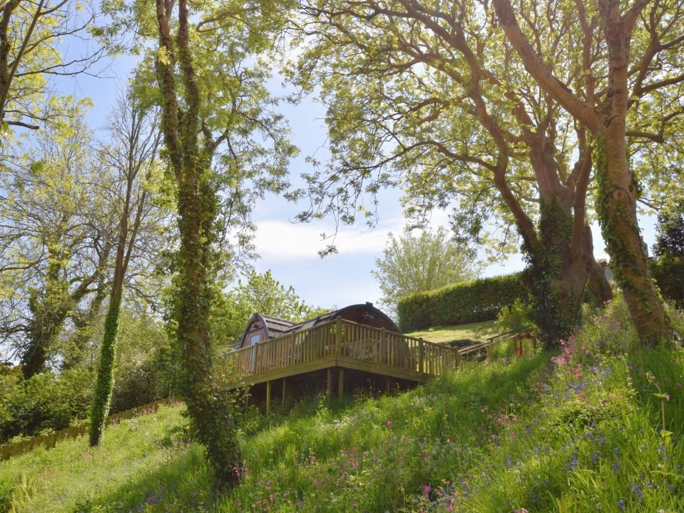 A wooden cabin with a deck, surrounded by lush greenery and flowering plants.