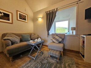 A cozy living room with a sofa, a small table with tea set, a window with curtains, and a television on the wall.