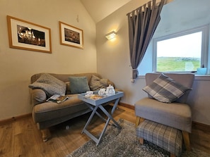 A cozy living room with a sofa, armchair, and a small table with a tray of tea and biscuits. There are two framed pictures on the wall and a window with curtains.