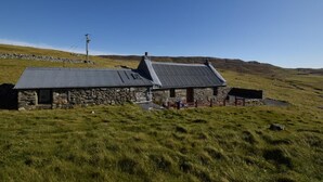 A stone house with a metal roof, surrounded by a grassy field and hills.