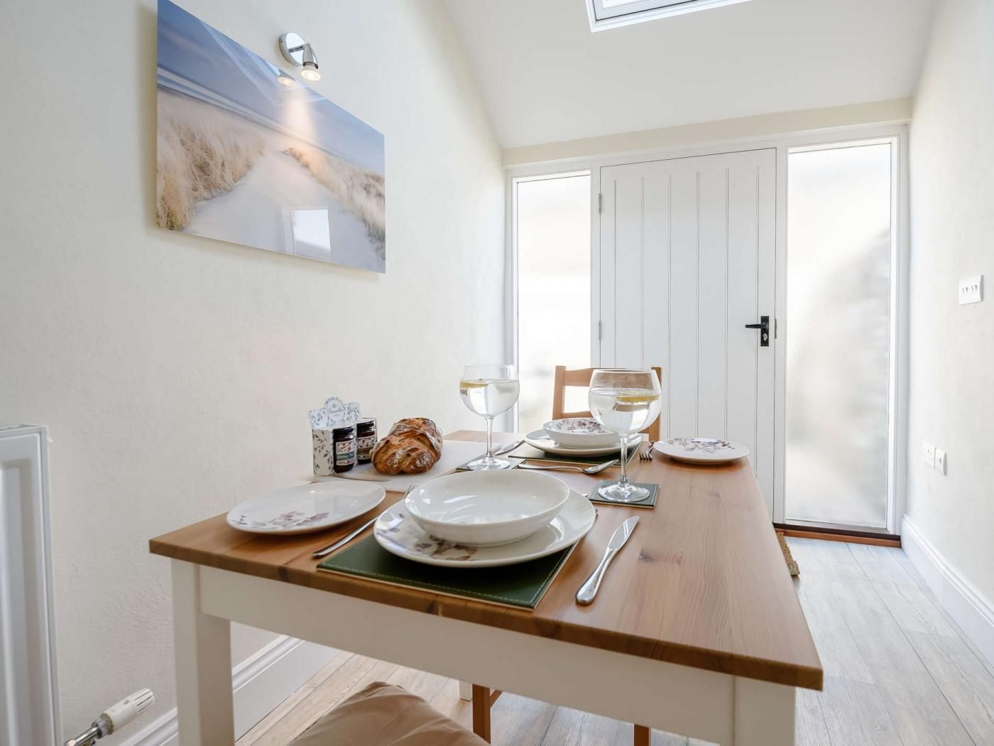 A dining area with a table set for two, a painting above the table, and a door in the background.
