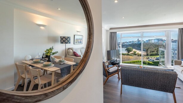 Een moderne woonkamer met een groot raam, een eethoek met een ronde tafel, en een spiegel die de kamer weerspiegelt.