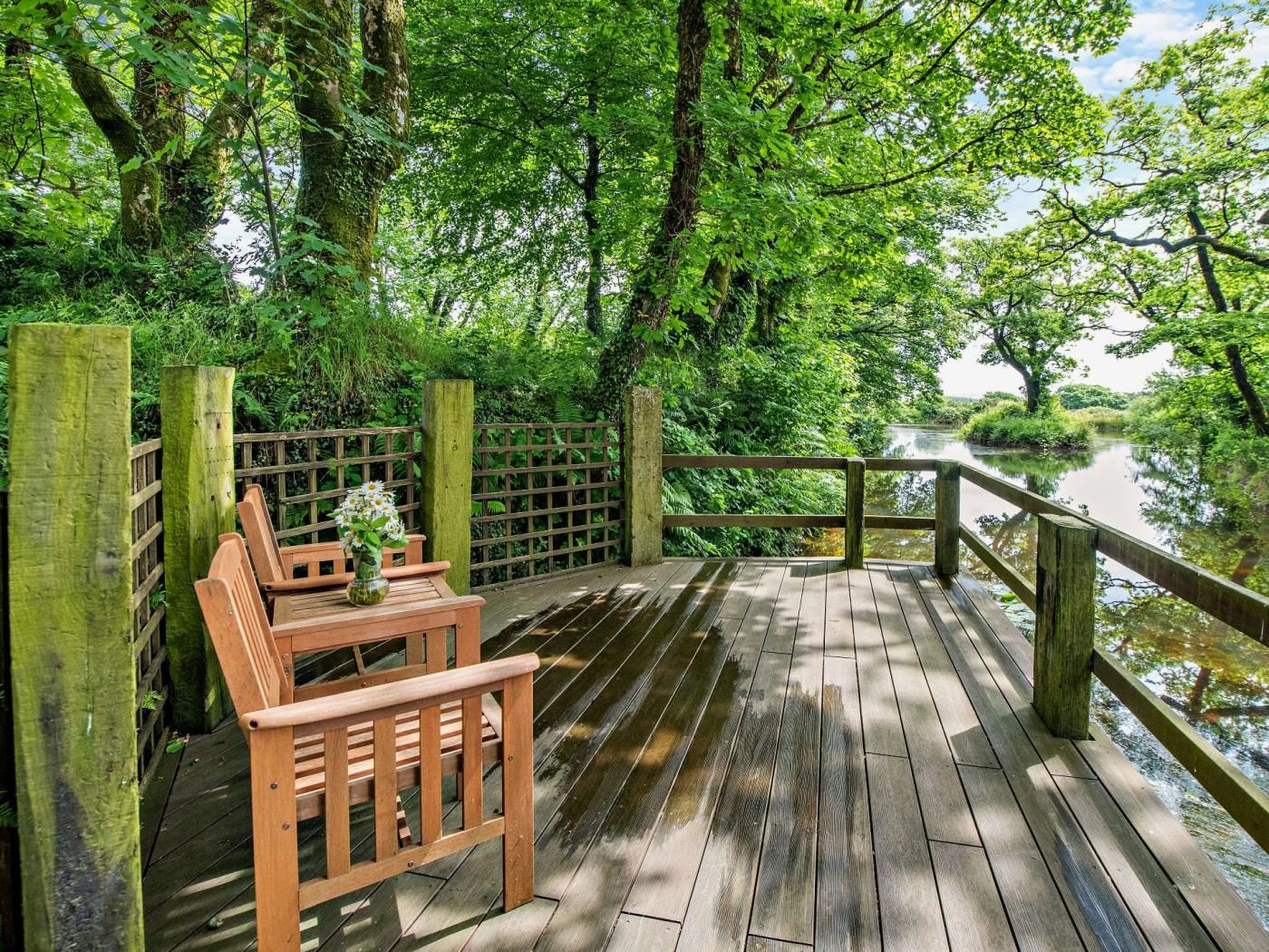 Une terrasse en bois avec un banc et une table, entourée d’une végétation luxuriante et bordée d’une rivière paisible.