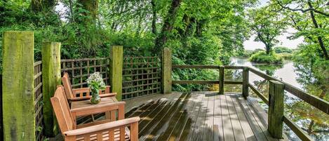 A wooden deck with a bench and a table, surrounded by lush greenery and a calm river.