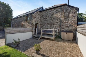 Ein Steinhaus mit einer Holzdielen-Terrasse, einer kleinen Außen-Sitzgelegenheit und einem Schotterweg.