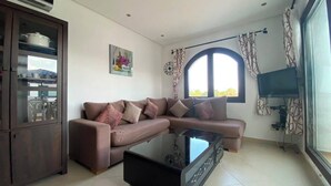 A living room with a sectional sofa, a glass coffee table, a TV, and a large window. - Serene 2-bedroom apartment in Cabo Negro with view of golf course (Cabo Negro)