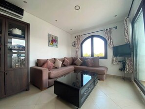 A living room with a sectional sofa, a glass coffee table, a TV, and a large window. - Serene 2-bedroom apartment in Cabo Negro with view of golf course (Cabo Negro)