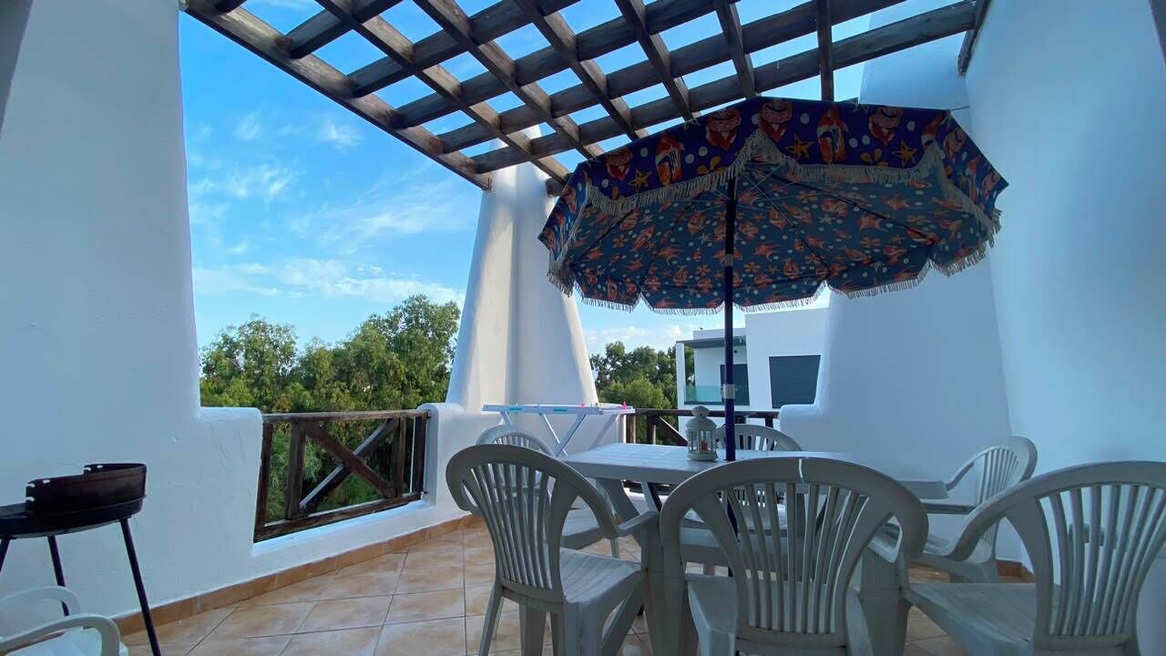 A covered outdoor area with a table and chairs, a patterned umbrella, and a view of trees.