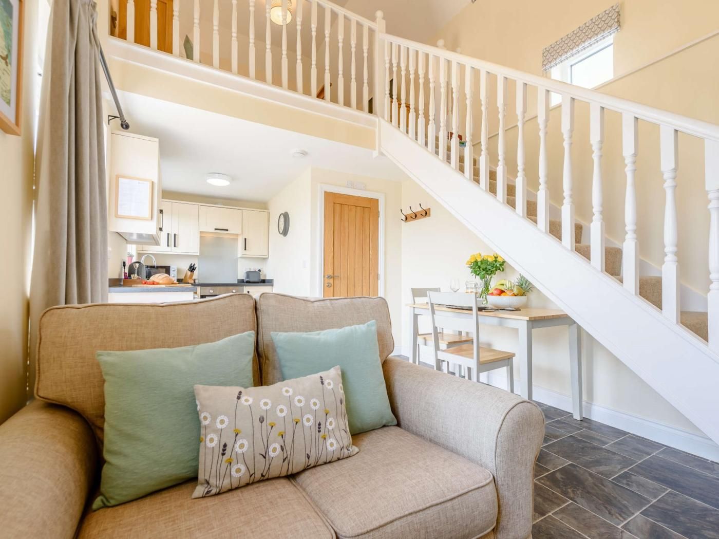 Un salon avec un canapé beige, deux coussins, un escalier avec des balustrades blanches et, au fond, une cuisine.