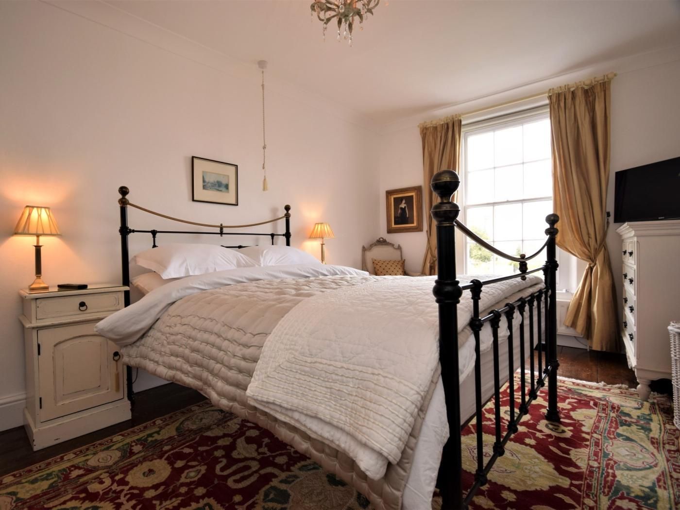 A bedroom with a four-poster bed, bedside lamps, a dresser, a television, and a large window with curtains.