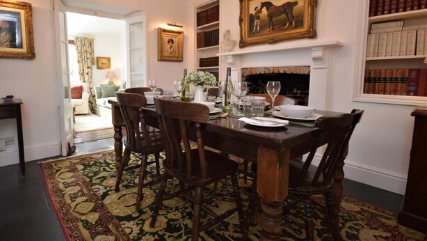 Une salle à manger avec une table en bois dressée pour six personnes, entourée de chaises, d’un lustre et d’une cheminée.