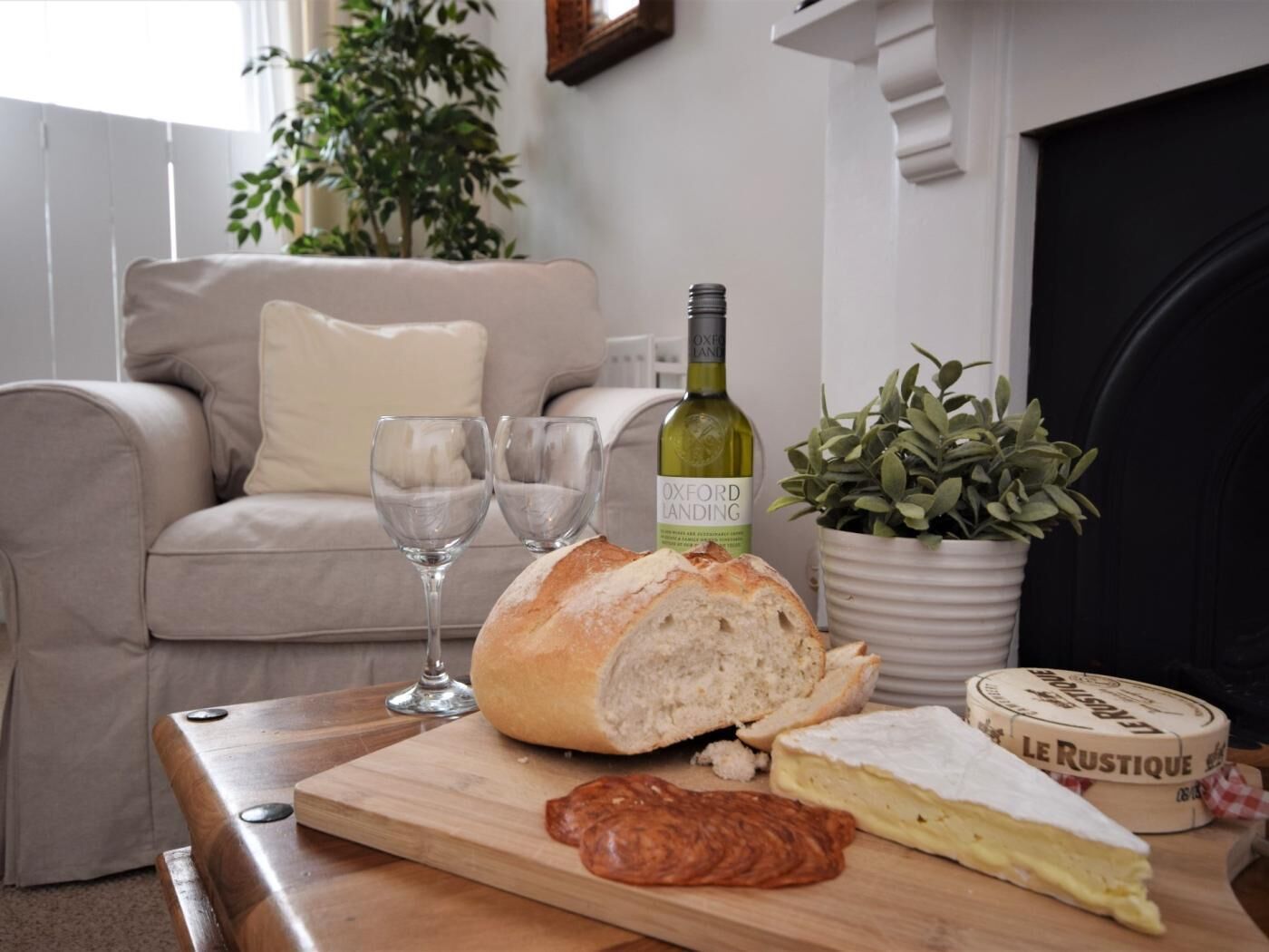 A living room with a sofa, a wooden table with a loaf of bread, two wine glasses, a bottle of wine, a potted plant, and a cheese wheel.