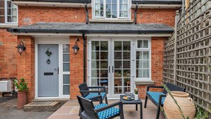 Une maison à deux étages, construite en briques, avec une porte d’entrée, deux fenêtres, un patio doté de mobilier d’extérieur et un grand réservoir d’eau.