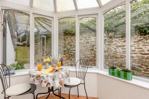 Une véranda avec une table dressée pour deux personnes, une chaise et une vue sur un mur de pierre et la verdure environnante.