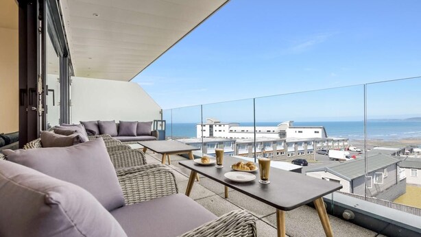 Ein Balkon mit Korbmöbeln, einem Tisch mit Tellern und Getränken, und Blick auf das Meer und Gebäude.