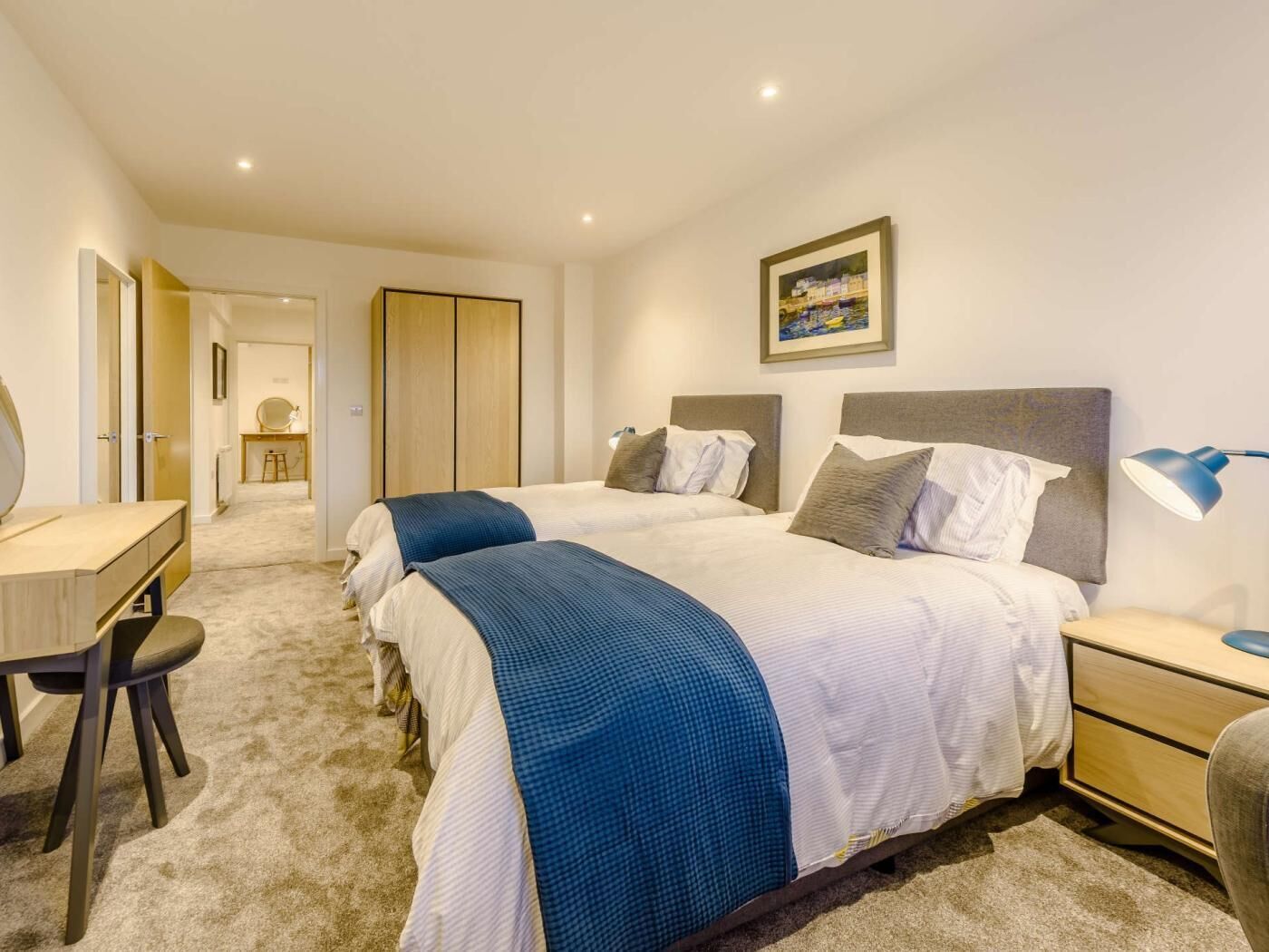 A hotel room with two beds, a desk, a chair, a bedside table, and a lamp.