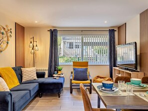 Ein modernes Wohnzimmer mit Sofa, Sessel, Esstisch, Fernseher und einem Fenster mit Jalousien.