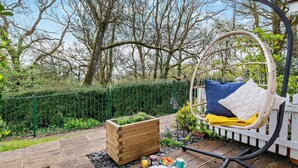 A wooden deck with a hanging chair, a wooden planter, and a table with a book and drinks. - 3 Bed in Bridport (oc-94692) (Fernhill)