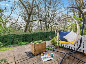 Eine Holzterrasse mit einem Hängesessel, einem Holzpflanzgefäß und einem Tisch mit Buch und Getränken.