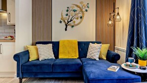 A modern living room with a blue sectional sofa, a decorative wall piece, and a dining area with a table and chairs. - 3 Bed in Bridport (oc-94692) (Fernhill)