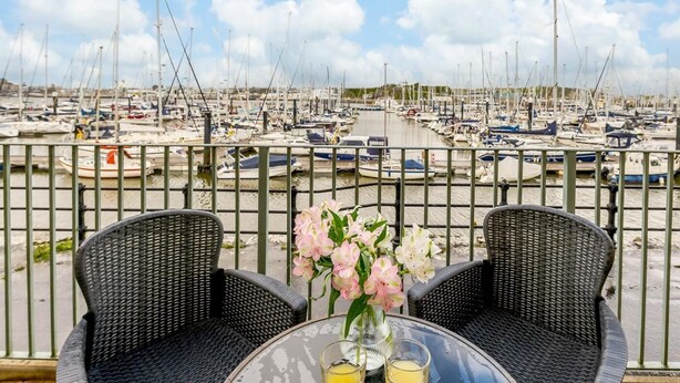Un balcon agrémenté de fauteuils et d’une table en osier, donnant sur une marina où sont amarrés de nombreux bateaux.