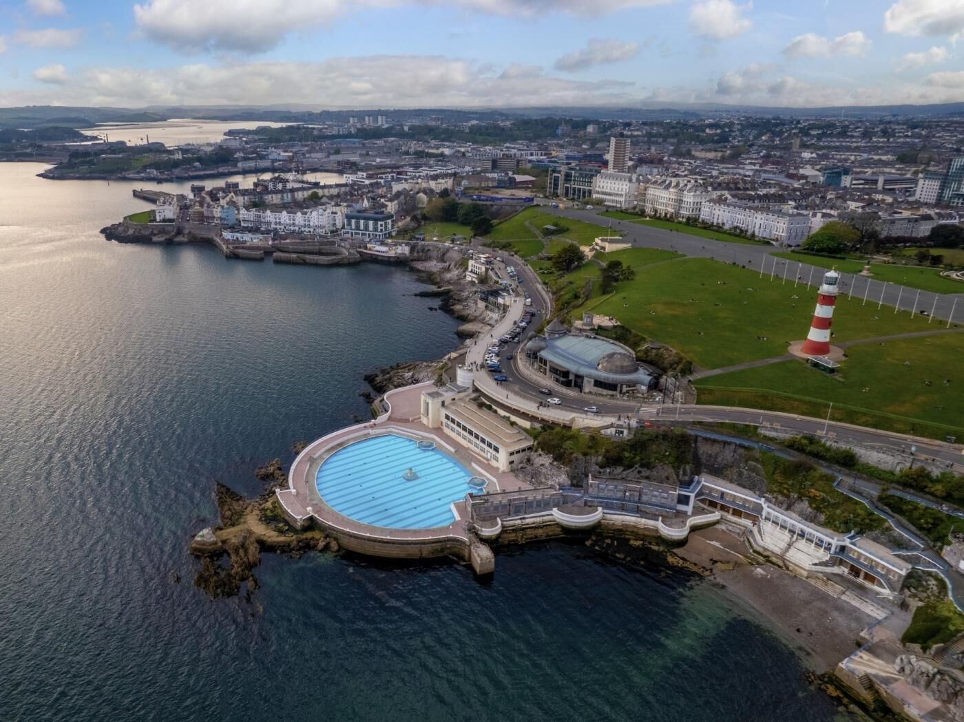 An aerial view of a coastal area with a swimming pool, a lighthouse, and a mix of residential and commercial buildings.