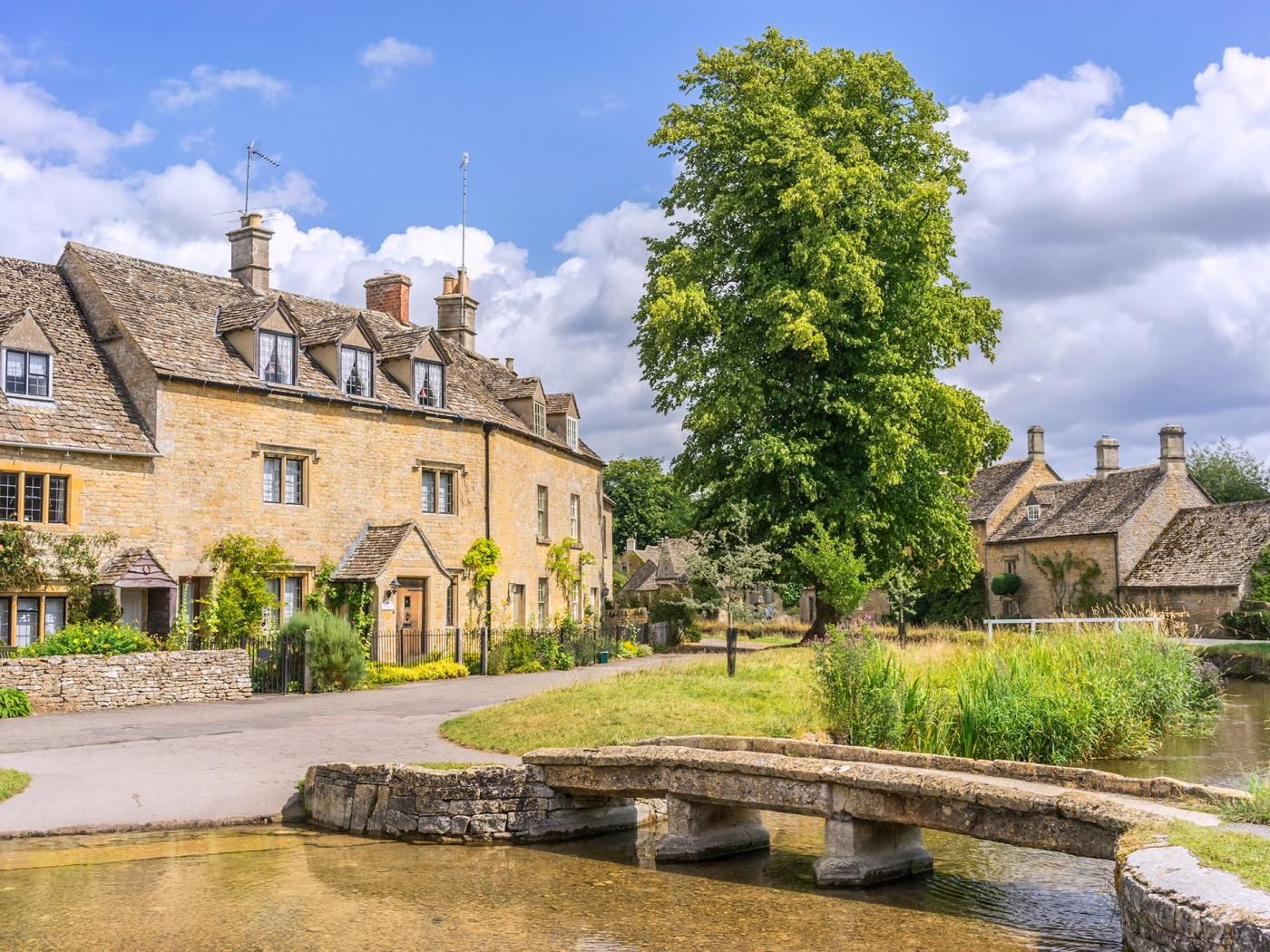 A stone building with multiple chimneys, a stone bridge over a body of water, and a large tree in the foreground.