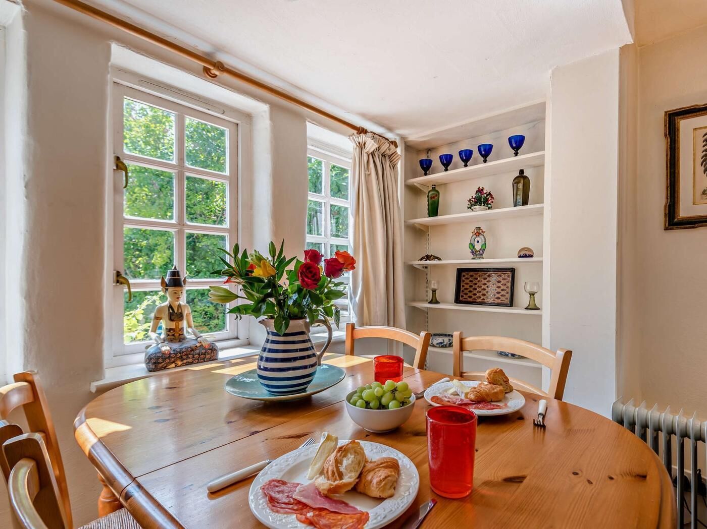 A dining area with a table set for two, a vase with flowers, and a view of greenery outside.