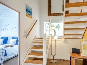 Ein modernes Interieur mit einer Treppe, einem Bett mit blauen Kissen, einem Schreibtisch mit einem Laptop und einem Fenster mit Blick auf Grünflächen.