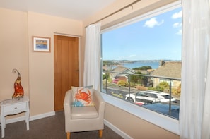 A room with a view of a body of water, a wooden door, a white chair with a colorful cushion, and a small table with a decorative item. - 4 Bed in Paignton (oc-56920) (Paignton)