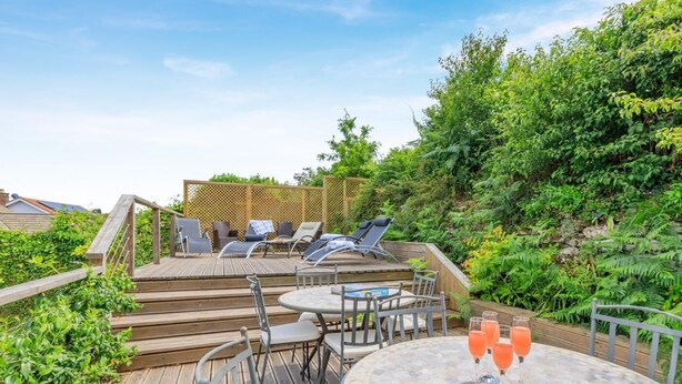 Une terrasse en bois avec des meubles d’extérieur, une table avec des boissons et, en arrière-plan, une végétation luxuriante.