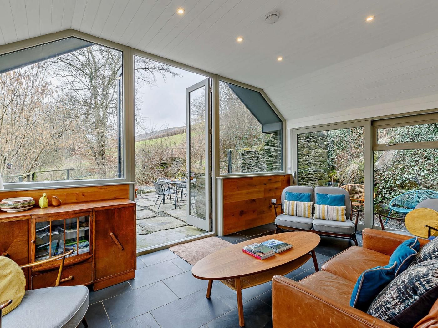 A spacious room with a large glass door leading to an outdoor area, a wooden cabinet with papers, a leather sofa with cushions, and a small round table with books.