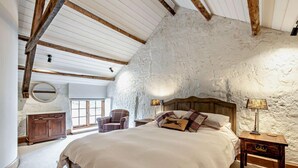 A bedroom with a stone wall, wooden beams, a bed, a chair, a nightstand, and a lamp. - 2 Bed in Exeter (oc-92739) (Longdown)