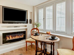A living room with a flat-screen TV, fireplace, dining table with chairs, and a window with blinds. - 1 Bed in Ilminster (oc-78353) (Ilminster)
