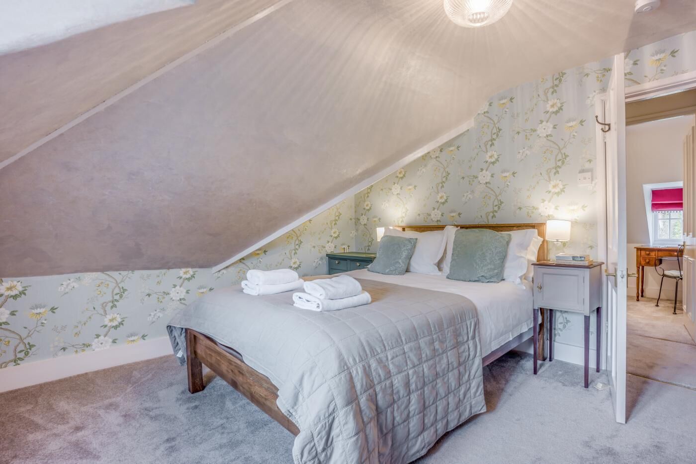 Et soveværelse med skrå væg, en seng med dyne, et natbord, en lampe og et skrivebord med stol.