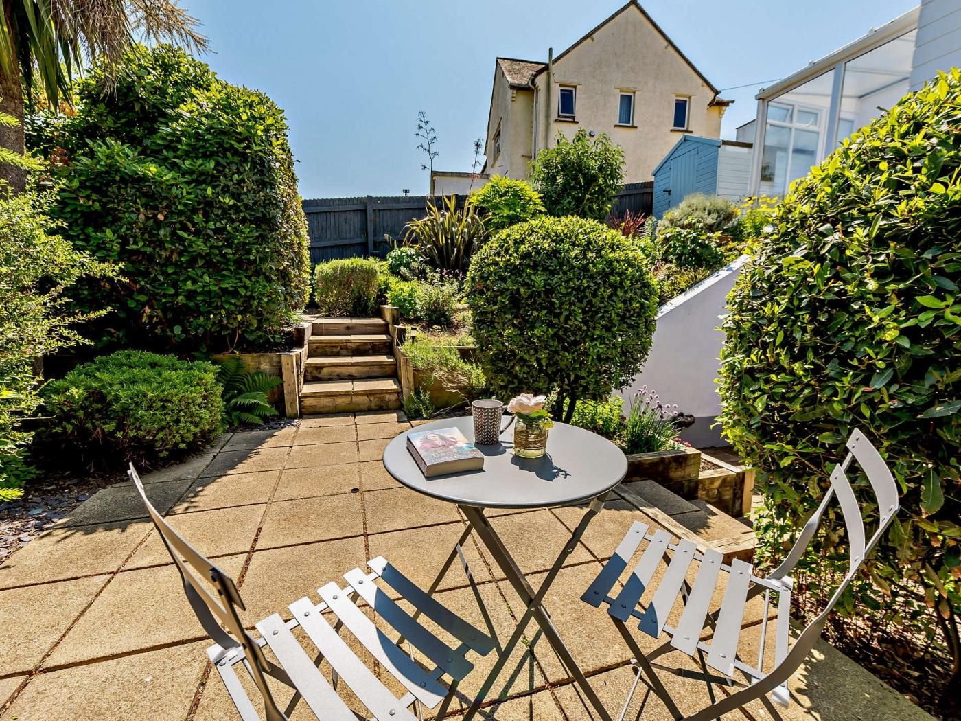 A patio with a round table and two chairs, surrounded by well-maintained hedges and a wooden staircase leading to a building.
