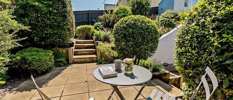 A patio with a round table and two chairs, surrounded by well-maintained hedges and a wooden staircase leading to a building.