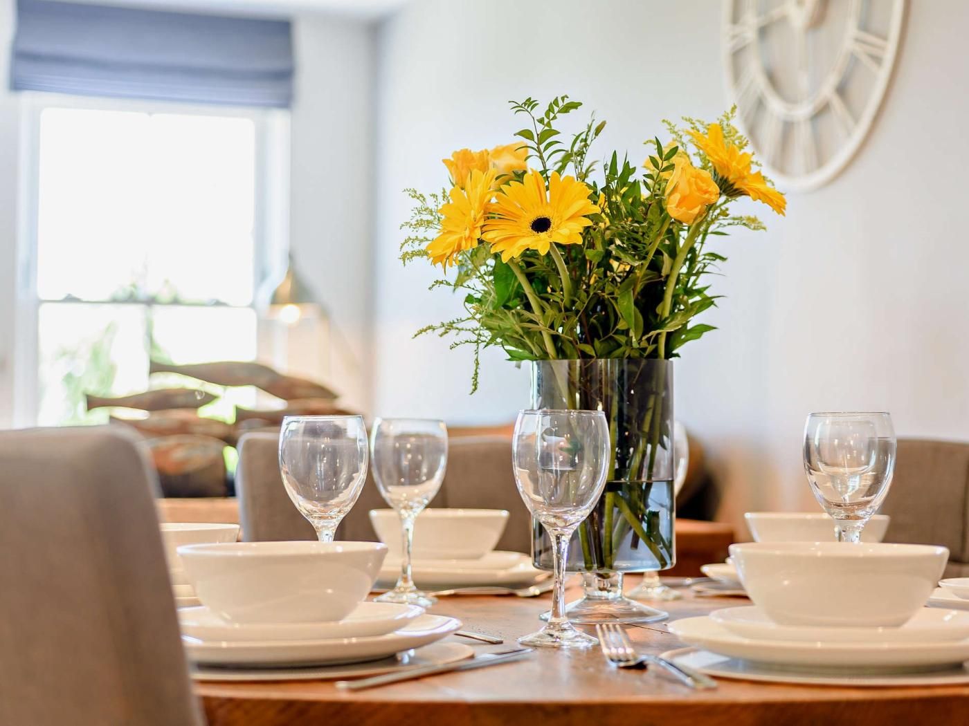 Een eettafel gedekt met witte borden, kommen en wijnglazen, met een vaas gele bloemen in het midden.