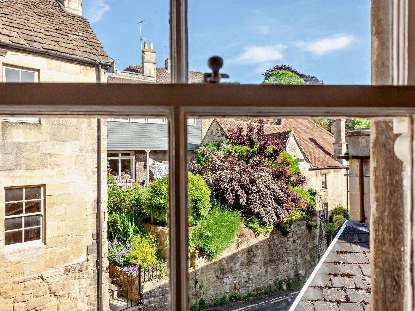 A stone building with a window, a narrow street, and a garden with various plants.